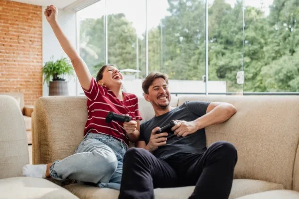 A man and woman are sitting on a light-colored sofa in a sunlit living room with large glass windows, laughing and holding video game controllers, with the woman raising her arm in celebration.
